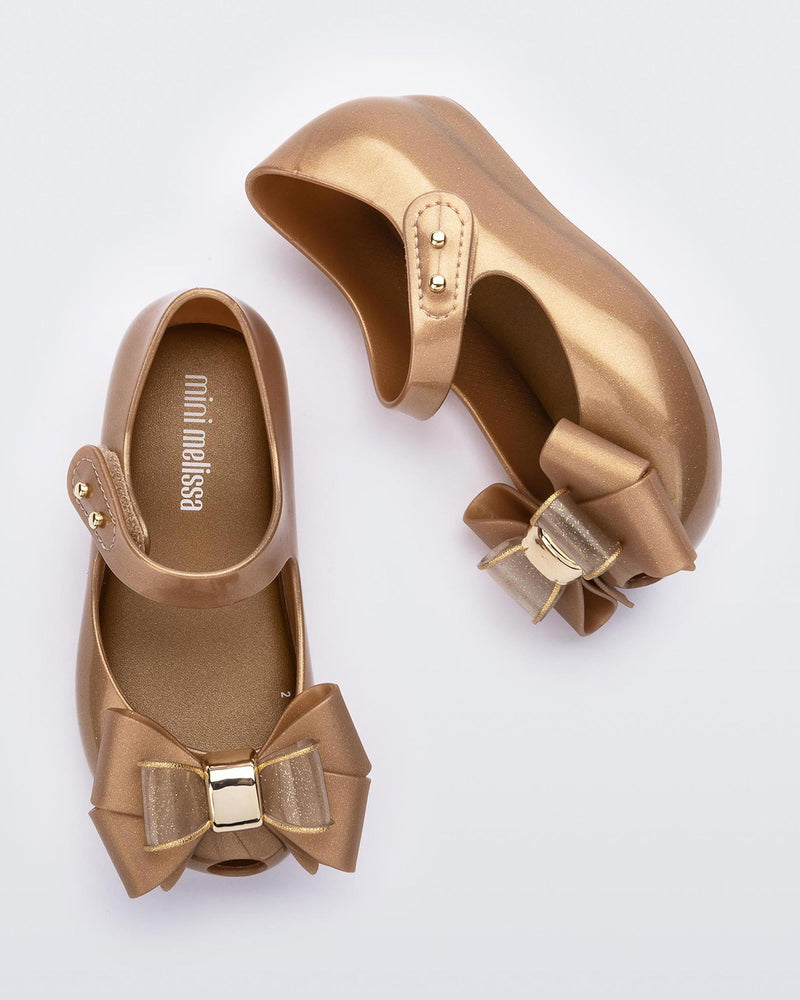 An angled top and side view of a pair of gold Mini Melissa Ultragirl Sweet flats with a top strap and a bow detail on the toe.
