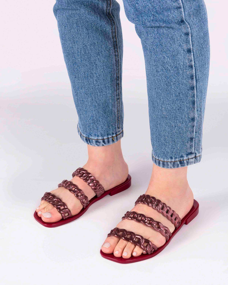 A model's legs wearing a pair of red Melissa Feel slides with three chain detail straps.
