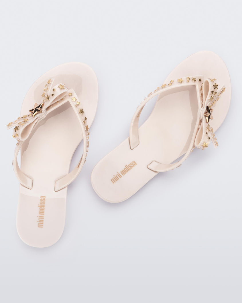 Top back view of a pair of beige Mini Melissa Harmonic Stars flip flops with a bow and gold star details on the straps.