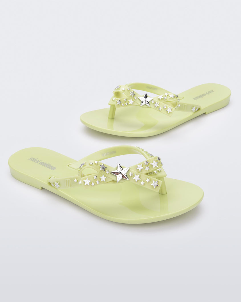 An angled front and side view of a pair of green Mini Melissa Harmonic Stars flip flops with a bow and silver star details on the straps.