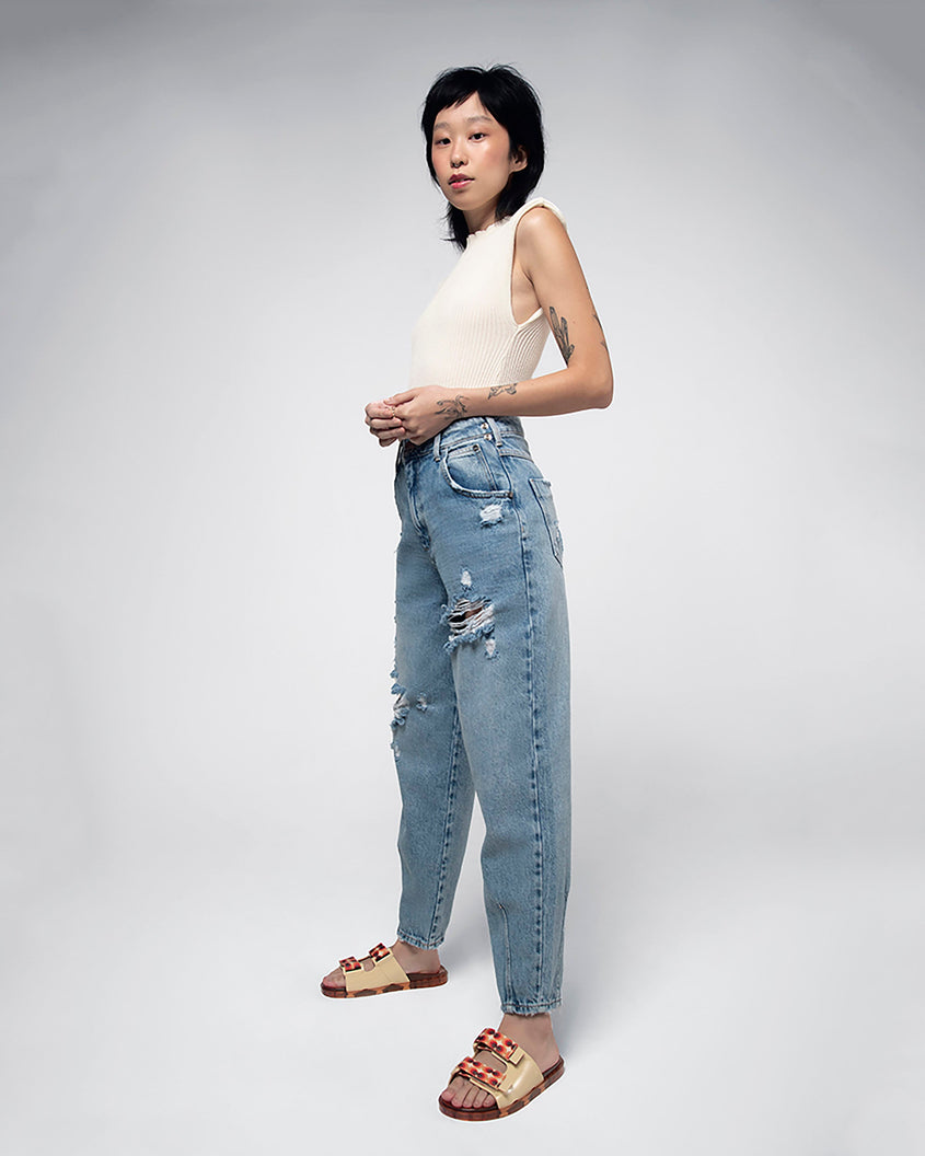 A model posing for a picture in jeans, a white tank top and a pair of yellow/orange Melissa Brave Wide Slides with a yellow base, red and orange circle print velcro straps and a translucent orange insole.