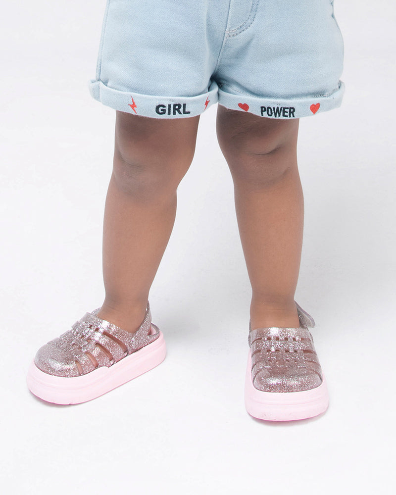 A young model's legs in denim shorts and a pair of pink/glitter Mini Melissa Sunday sandals with a glitter base, closed toe front and pink sole.