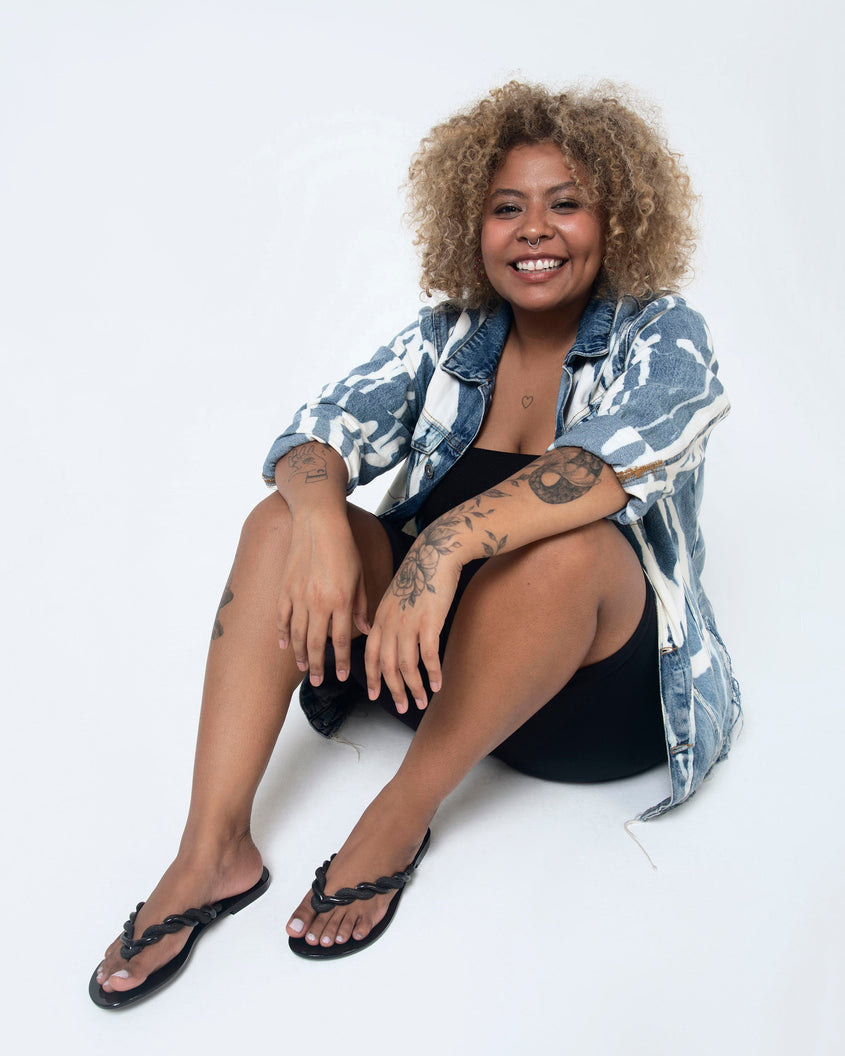 A model sitting on the ground with her legs crossed, posing for a picture in a pair of Black/Gold Melissa Louise Flip Flops with glitter twisted rope like straps.