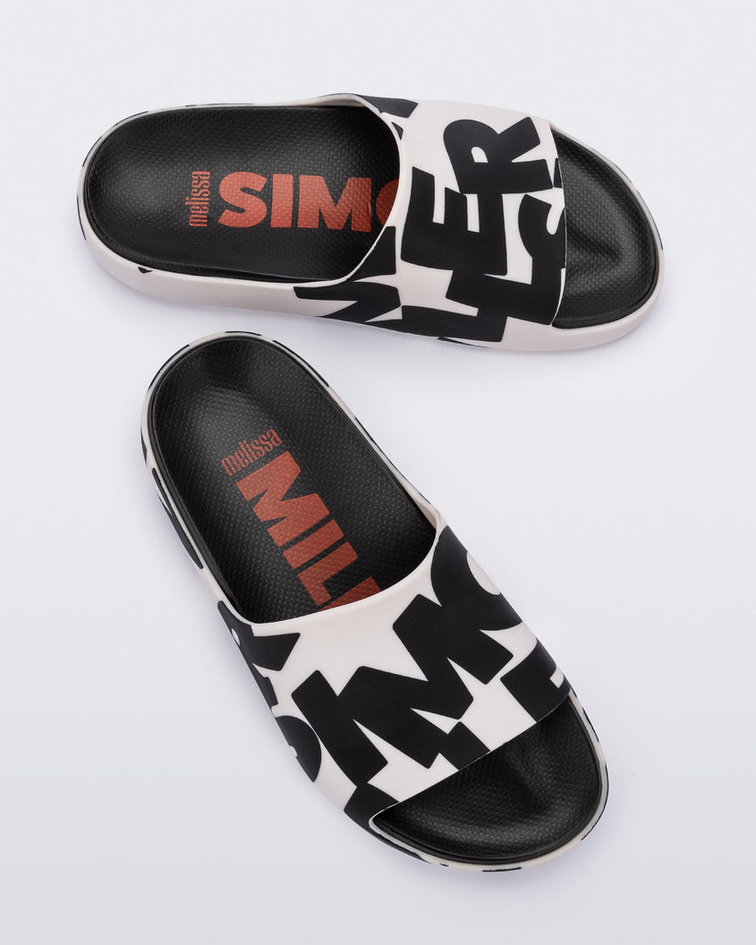 A top and side view of a pair of White Logo Scramble Melissa Cloud slides with a black simon miller letter scrambled text on a white base base and a black insole reading in red