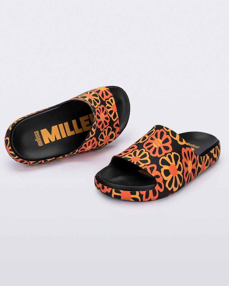 An angled top and front view of a pair of multicolor matte mosaic flower Melissa Cloud slides with a black base and an orange and yellow flower pattern, with a black insole reading in orange "Melissa Miller" and "Melissa Simon" on different shoes