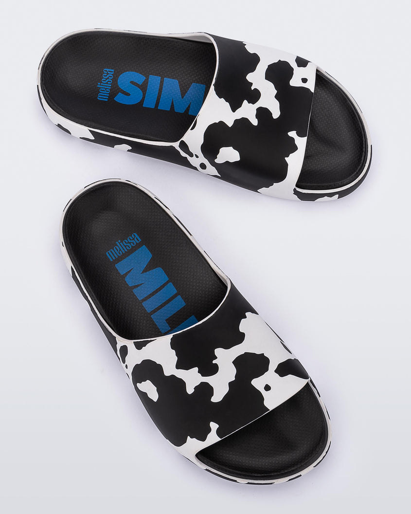 An angled top and side view of a pair of black and white cow print Melissa Cloud slides with a black insole reading in blue