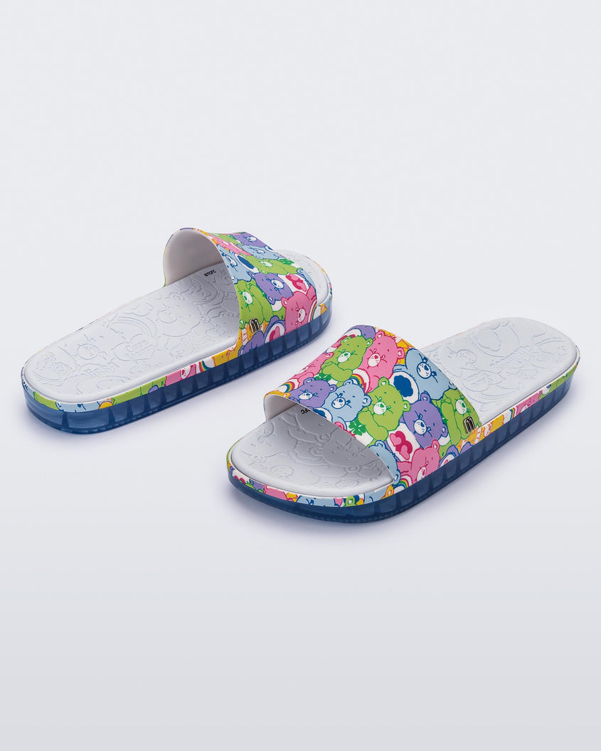 An angled front and side view of a pair of blue Melissa Beach slides with a pattern of lilac, green, pink and blue colored care bears on the top strap, and a care bear patterend gray insole.
