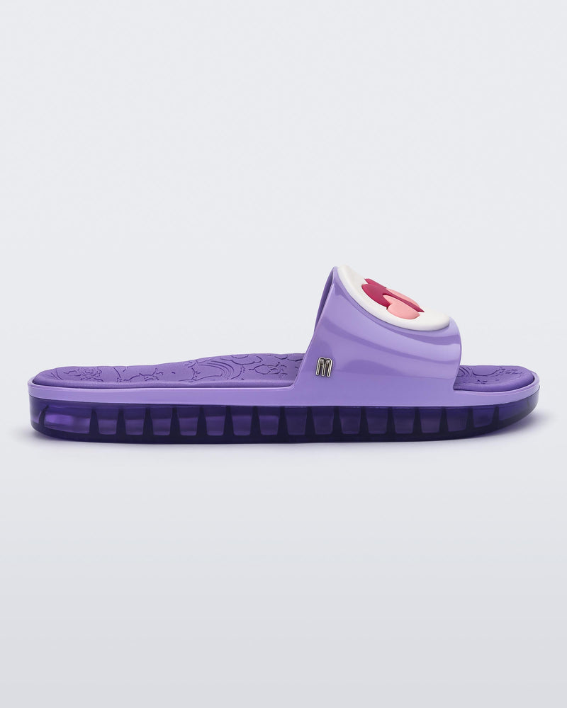 Side view of a lilac Melissa Beach slide with a silver "m" on the side, 3 pink hearts on a white circle on the top strap, and a care bear patterend lilac insole.