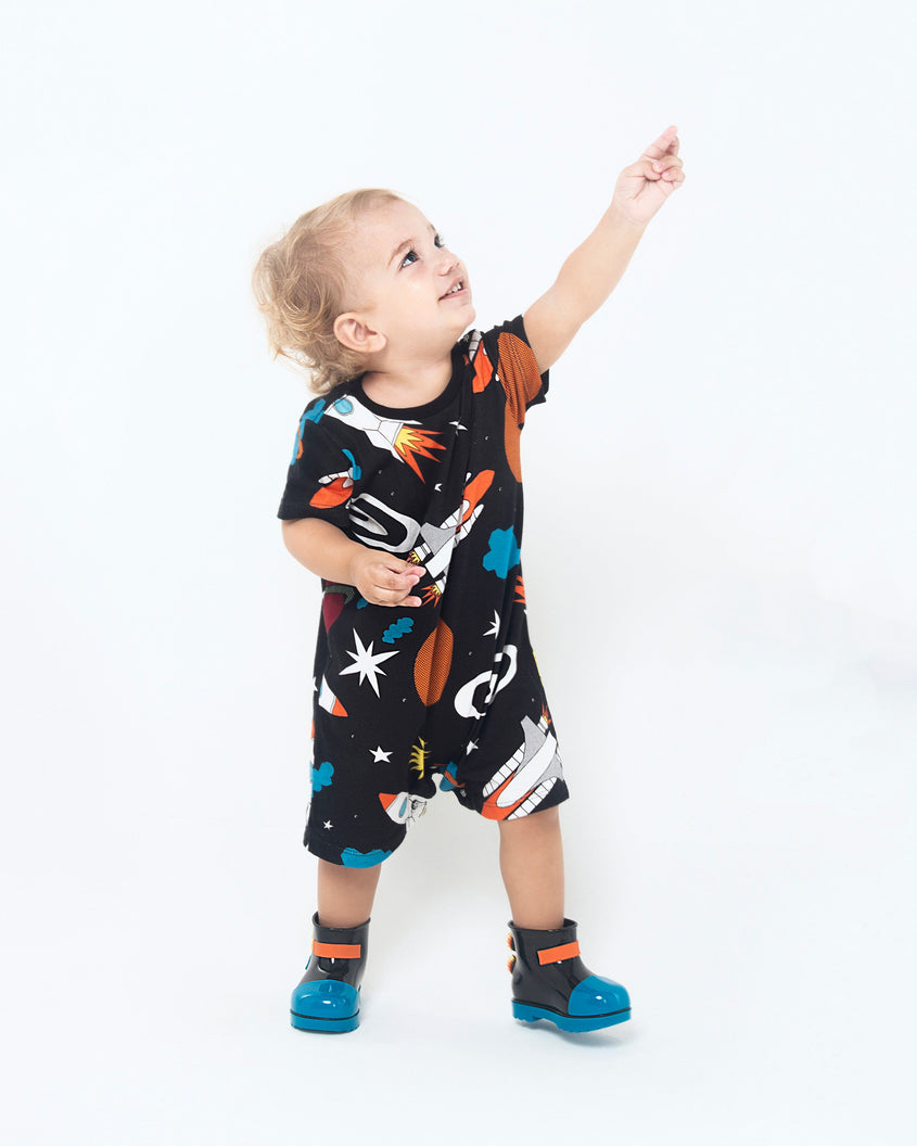 A baby model posing for a picture in a space themed onesie and a pair of blue/black Mini Melissa Rain Boots with a black base, blue toe, blue sole, and a planet, star and fire detail on the side and back.