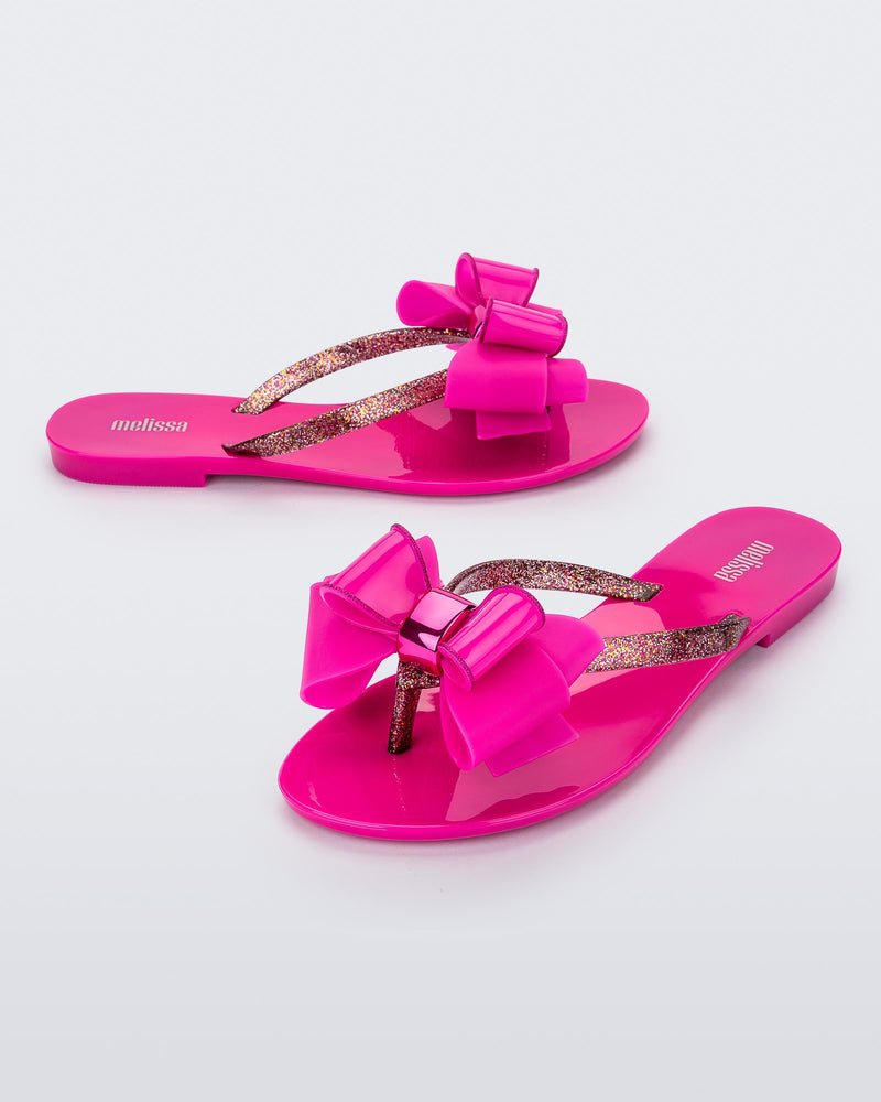 An angled side and front view of a pair of dark pink Melissa Harmonic Sweet flip flops with a pink bow and multicolor glitter straps.