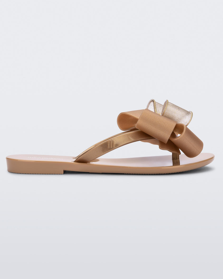 Side view of a beige Mini Melissa Harmonic Sweet flip flop with a beige and clear gold glitter bow with a gold buckle.