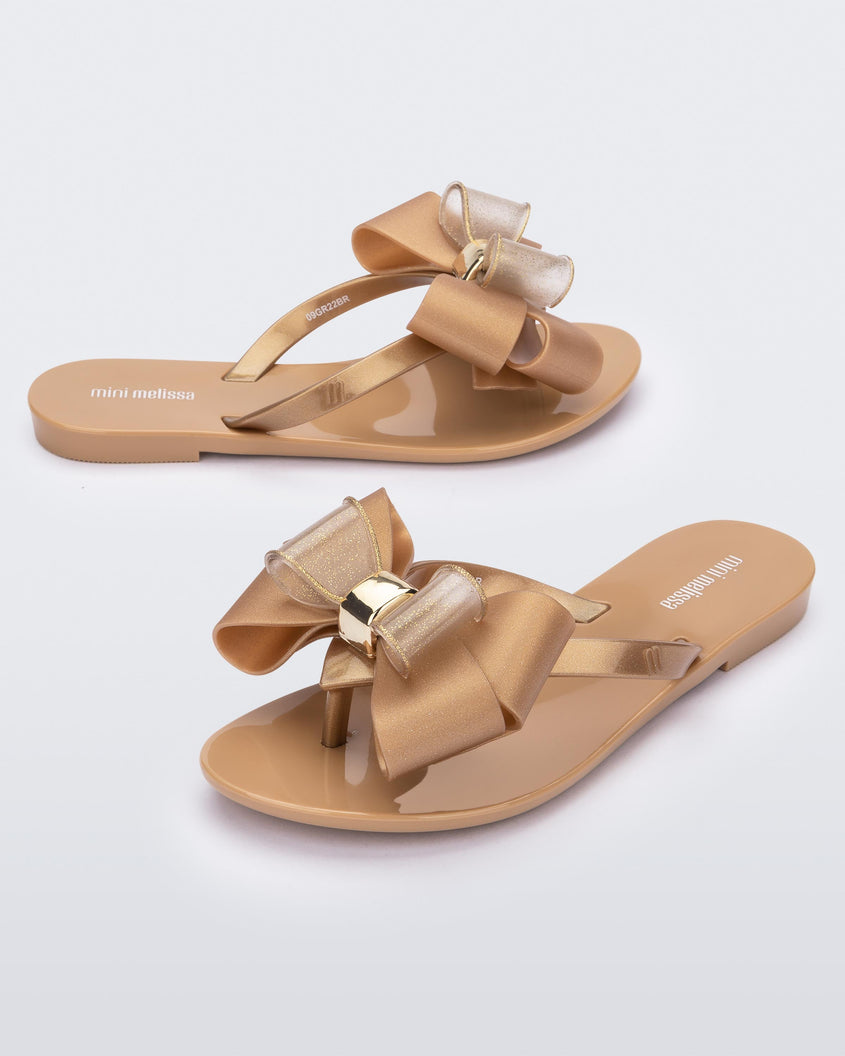 An angled side and front view of a pair of beige Mini Melissa Harmonic Sweet flip flops with a beige and clear gold glitter bow with a gold buckle.