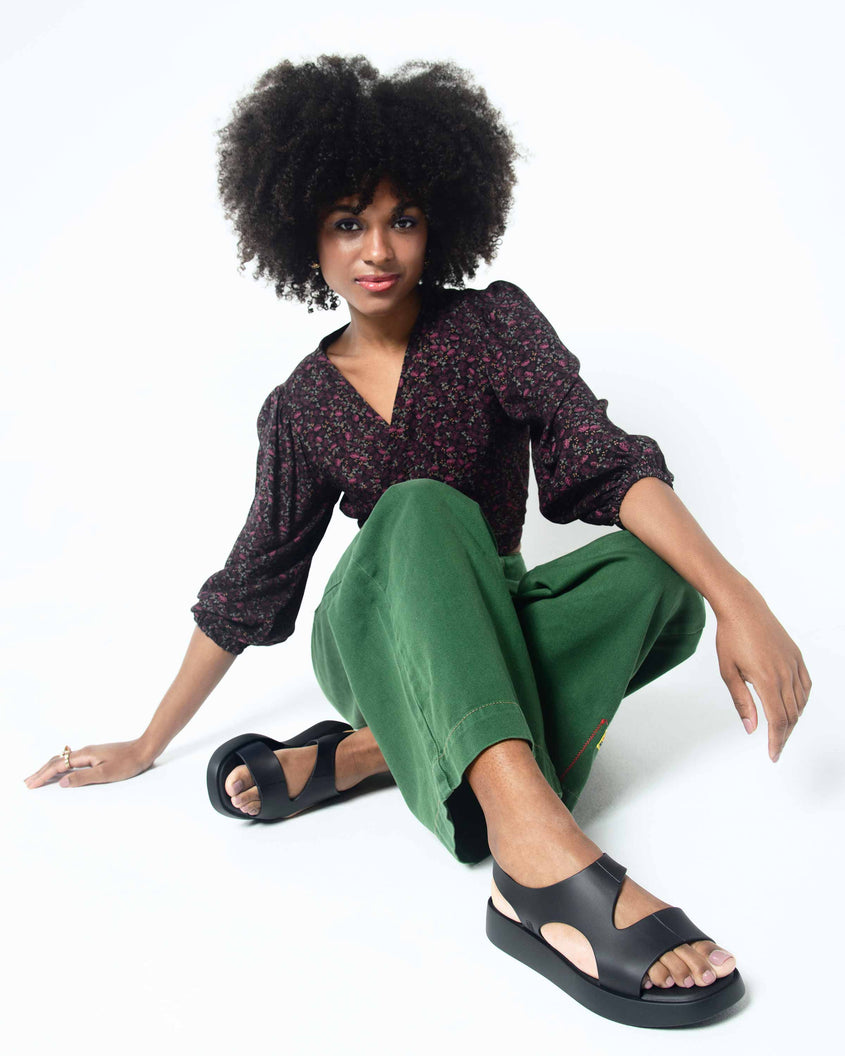 A model sitting on the ground with her legs crossed posing for a photo wearing a pair of black Melissa Franny sandals with straps.