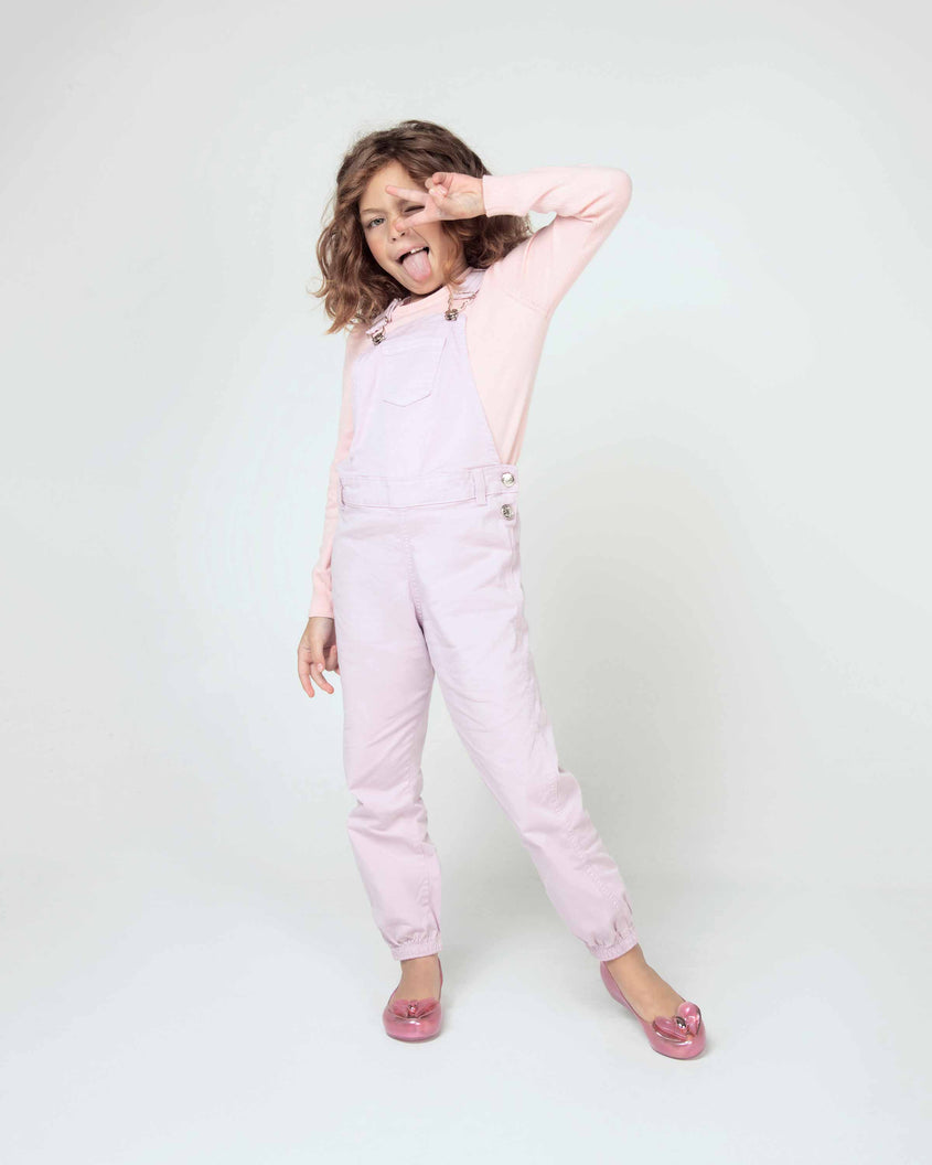 A model posing for a picture in a pink top, overalls and a pair of a pearly pink Mini Melissa Ultragirl flats with a heart buckle bow detail on the toe.