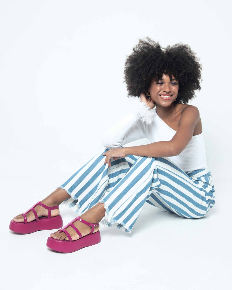 A model sitting on the ground, posing for a picture in a white top, blue and white striped bottoms and a pair of pink Melissa Caribe High Platform sandals with two pink front straps joined by a vertical strap intersecting the ankle strap and a black sole.