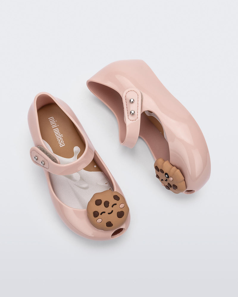 Top and side view of a pair of Mini Melissa Ultragirl Candy ballet flats in Pink colorway. The shoe has a single strap across the top of the foot with a double snap closure. The product also has a 3D chocolate chip cookie applique on the top fo the toe. The cookie applicque is a light brown color with darker brown colored chips.