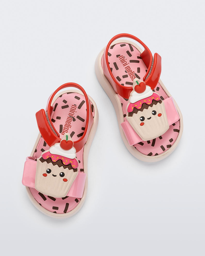 Top view of a pink/red/yellow Mini Melissa Jump Candy sandals with a print of sprinkles on the insole and a plastisol cupcake on top of the straps.