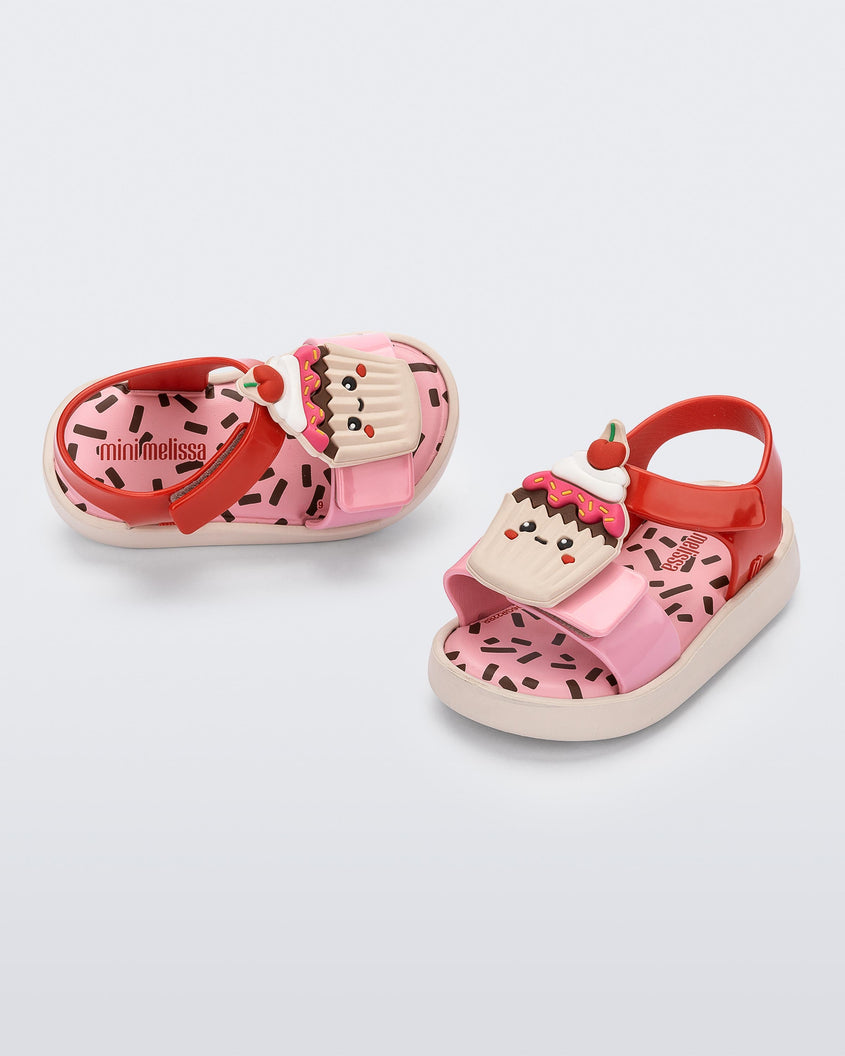 An angled top and side view of a pair of pink/red/yellow Mini Melissa Jump Candy sandal with a print of sprinkles on the insole and a plastisol cupcake on top of the straps.