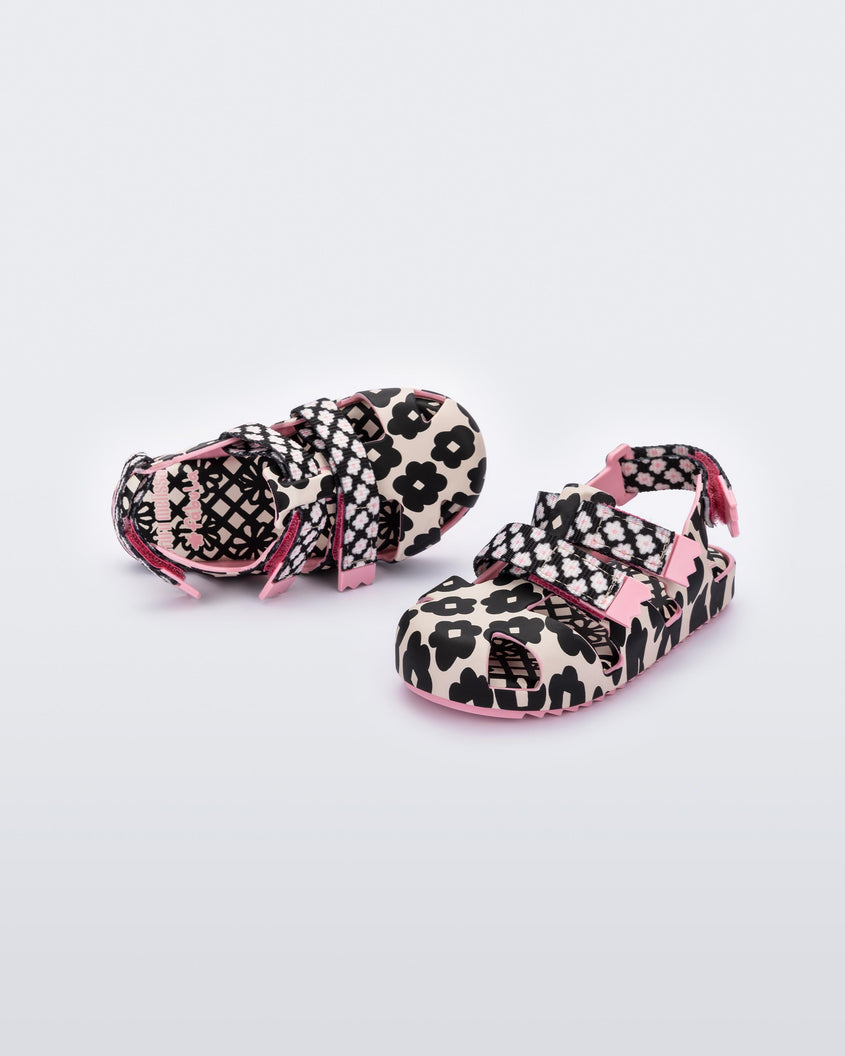 An angled front and top view of a pair of pink/black sandals Mini Melissa Yoyo with a pink and black floral base, two velcro front straps and a back ankle strap.