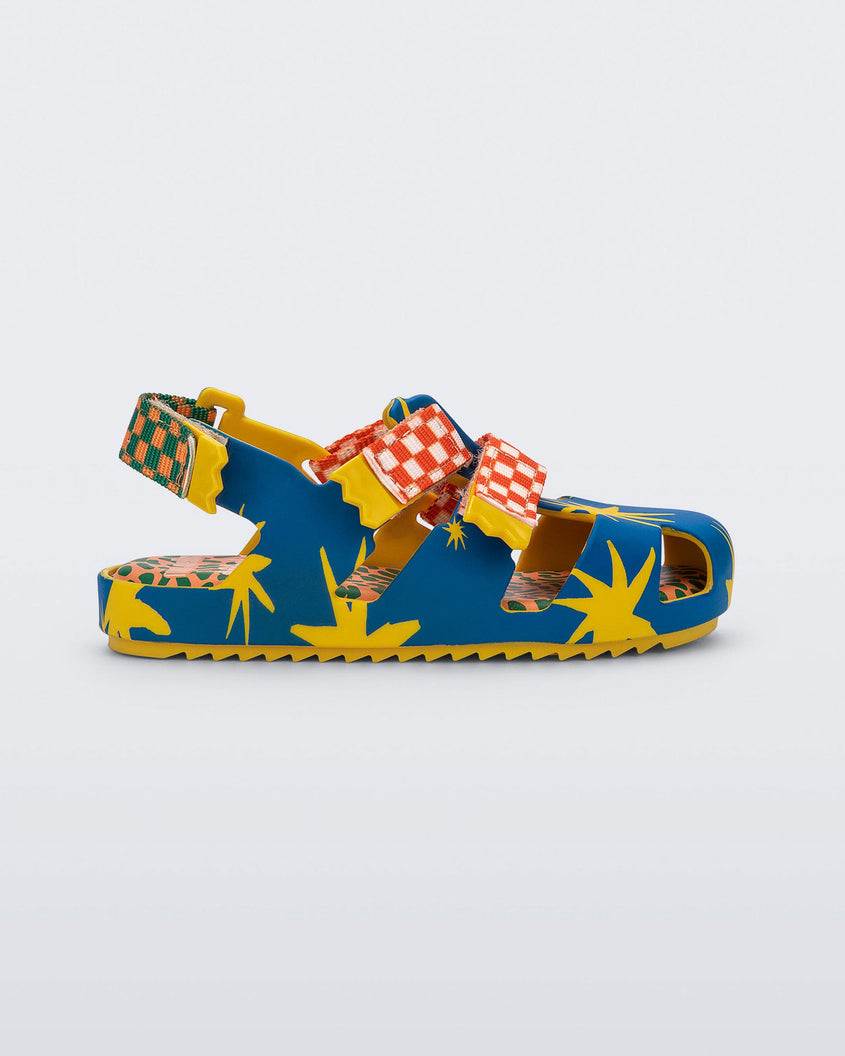 Side view of a yellow/blue Mini Melissa Yoyo sandal with a yellow and blue patterned base, with two red and white velcro front straps and an orange and green ankle strap.