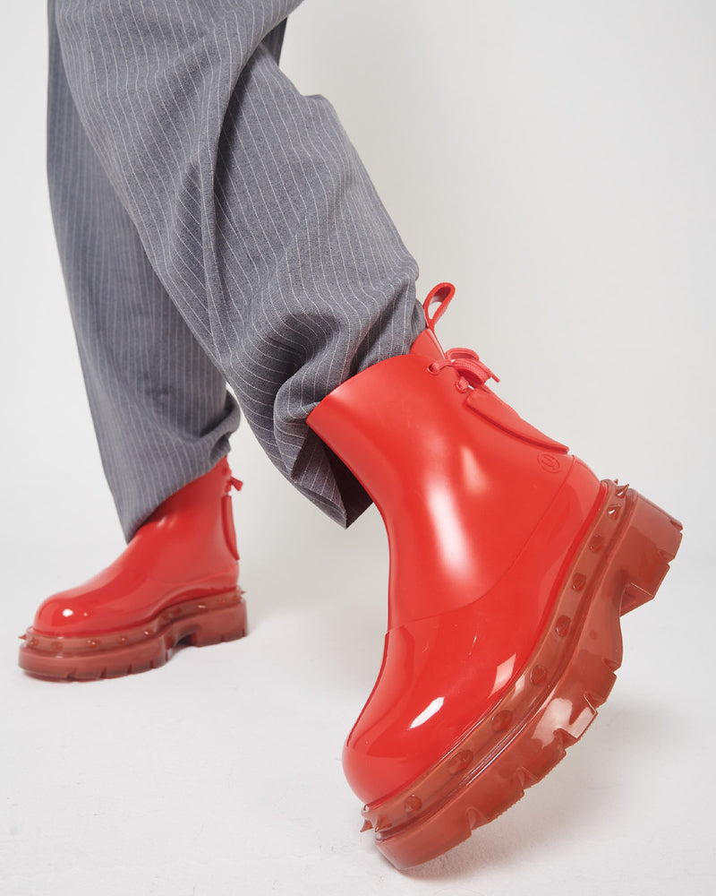 A model's legs in gray pants and a pair of red Melissa Spikes Boots with spike details around the sole and laces in the back.