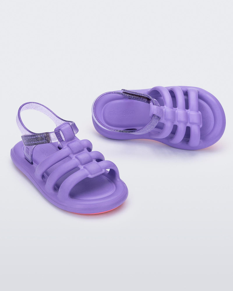 An angled front and side view of a pair of lilac Mini Melissa Freesherman sandals with lilac and clear glitter straps.