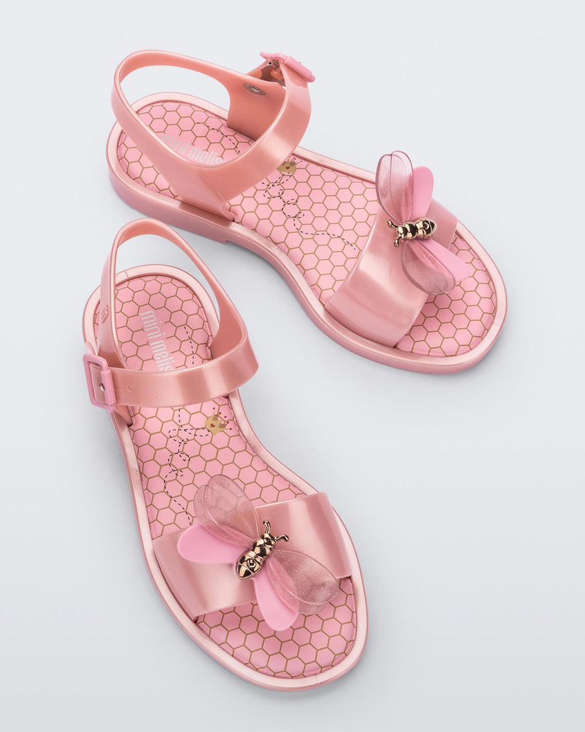 An angled top view of a pair of pearly pink Mini Melissa Mar Bugs sandal with a honeycomb pattern insole and a pink bug detail with a gold buckle on the front.