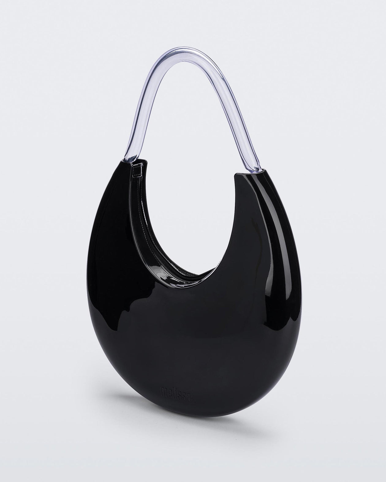Angled view of the Melissa Moon Handbag. The body of the bag is crescent shaped and is black with a top zipper closer. There is a short shoulder handle that is clear / transparent material.