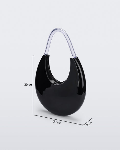 Angled view of the Melissa Moon Handbag. The body of the bag is crescent shaped and is black with a top zipper closer. There is a short shoulder handle that is clear / transparent material. This image contains dimension details. The bag is 29 cm in length, 30 cm in height (excludes handle height) and 6 cm width.
