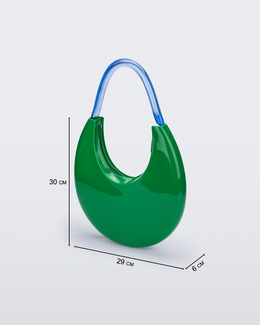 An angled front view of a Green/Blue Melissa Moon Bag with a crescent shaped green bag base and a blue beige top strap. The image also shows the dimensions of the bag which reads 30 centermeters high, 29 centermeters wide and 6 centermeters deep.