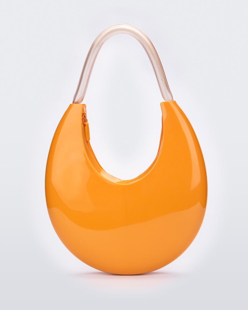 A zoomed in front view of a orange/beige Melissa Moon Bag with a crescent shaped orange bag base and a clear beige top strap.