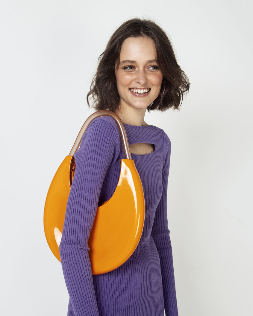 A zoomed in shot of a model posing for a picture in a purple dress, and showing an orange/beige Melissa Moon Bag with a crescent shaped orange bag base and a clear beige top strap on her shoulder.