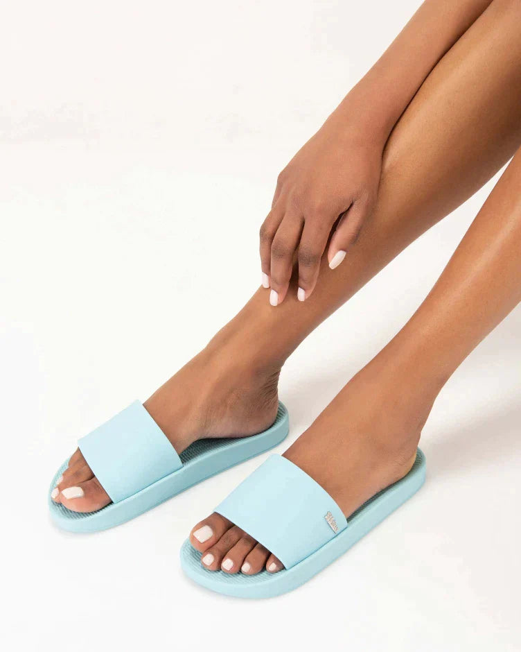 A model's legs wearing a pair of light blue Melissa Sun Sunset slides with a