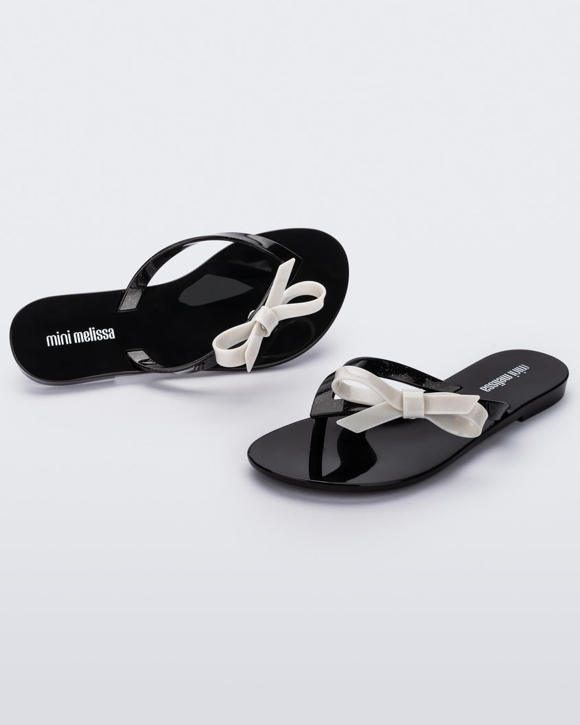 An angled front and top view of a pair of black/glitter white Mini Melissa Harmonic Sweet flip flops with a black base, black glitter straps and a white bow on top.