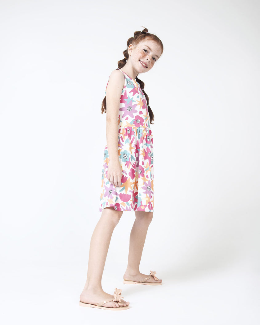 A model posing for a picture in a white patterned dress and a pair of beige Mini Melissa Slim flip flops with a lace like bow detail.
