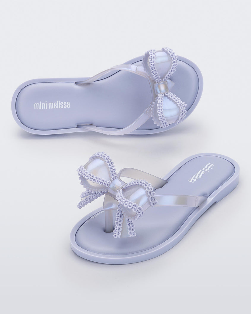 An angled front and side view of a pair of lilac Mini Melissa Slim flip flops with a lace like bow detail on the front strap.