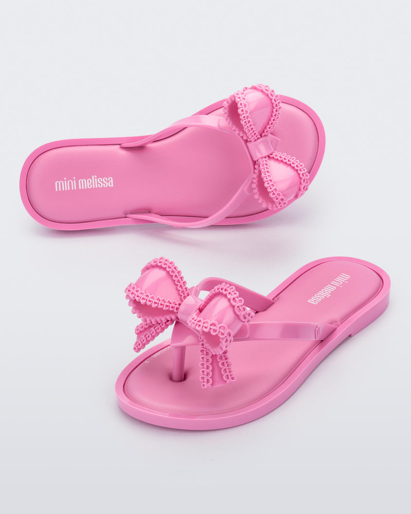 An angled front and view of a pair of pink Mini Melissa Slim flip flops with a lace like bow detail on the front strap.