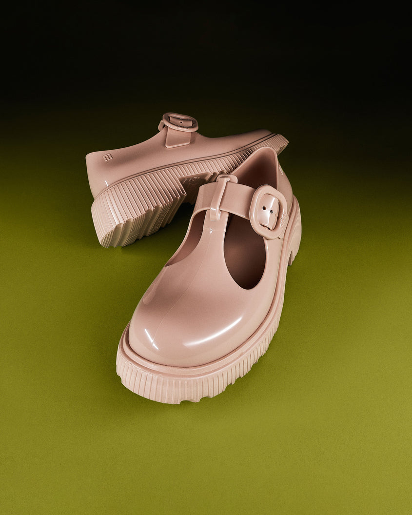 An angled front and side view of a pair of beige Melissa Jackie loafers with two cut outs and a buckle detail strap on a green background.