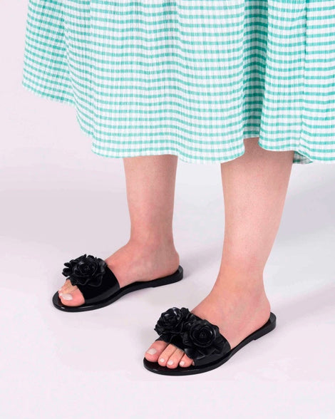A model's legs wearing green gingham printed dress and a pair of black Melissa Babe Garden slides with two flowers on the front strap.