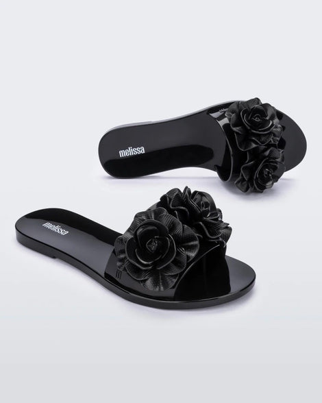 An angled side and top view of a pair of black Melissa Babe Garden slides with two flowers on the front strap.