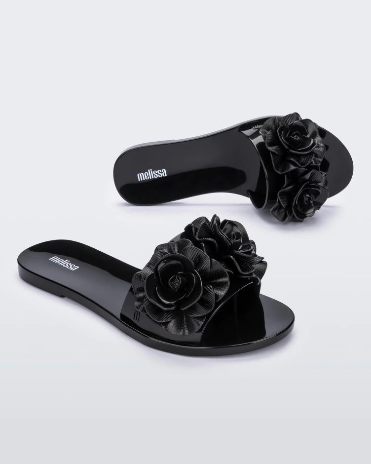 An angled side and top view of a pair of black Melissa Babe Garden slides with two flowers on the front strap.