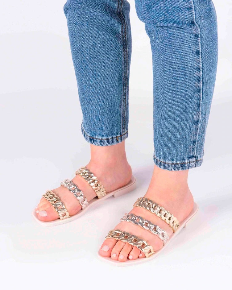 A model's legs wearing a pair of white Melissa Feel slides with three silver and gold chain detail straps.