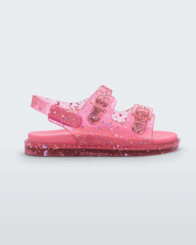 Side view of a pink Mini Melissa Wide Sandal with a clear pink base with a splatter paint detail, two buckles at the top, a velcro ankle strap and a pink insole.