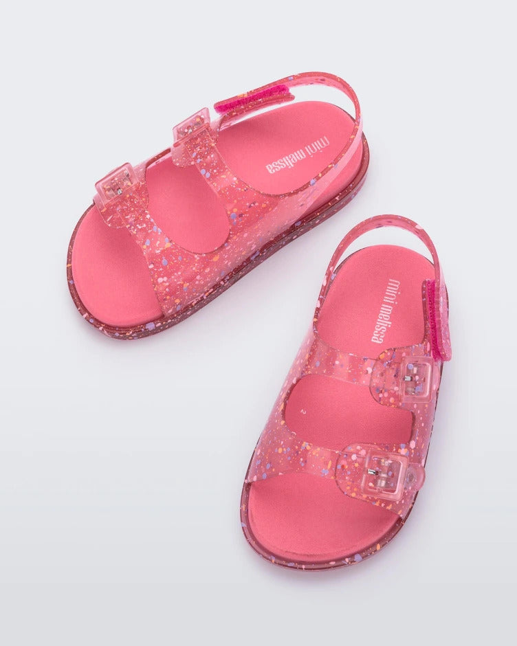 Top view of a pair of pink Mini Melissa Wide Sandals with a clear pink base with a splatter paint detail, two buckles at the top, a velcro ankle strap and a pink insole.