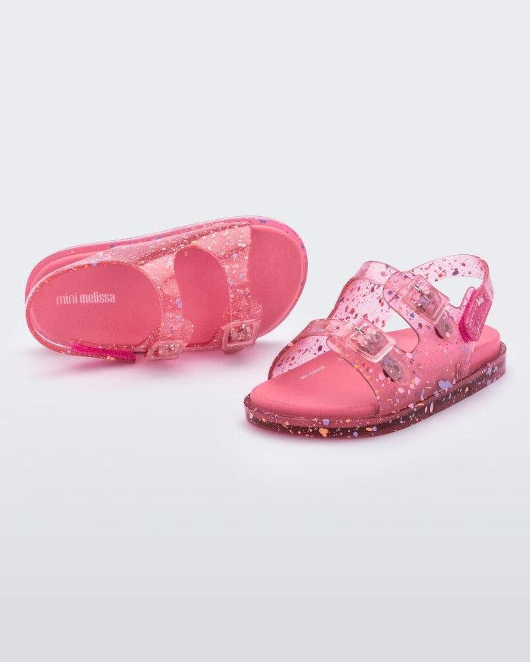 A front and top view of a pair of pink Mini Melissa Wide Sandals with a clear pink base with a splatter paint detail, two buckles at the top, a velcro ankle strap and a pink insole.