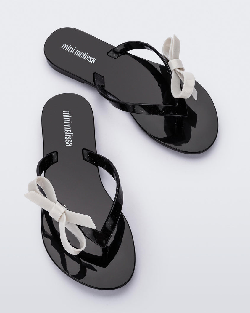 Top view of a pair of black/glitter white Mini Melissa Harmonic Sweet flip flops with a black base, black glitter straps and a white bow on top.