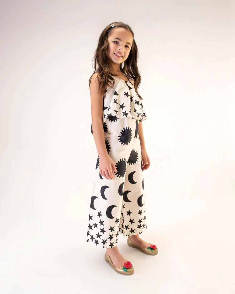 A model in a black and white patterned jumpsuit, wearing a pair of gold Mini Melissa Sweet Love Princess flats, with a gold base, Belle in script on the side, a rose detail on the toe, and a drawing of Belle of Beauty and the Beast on the sole.