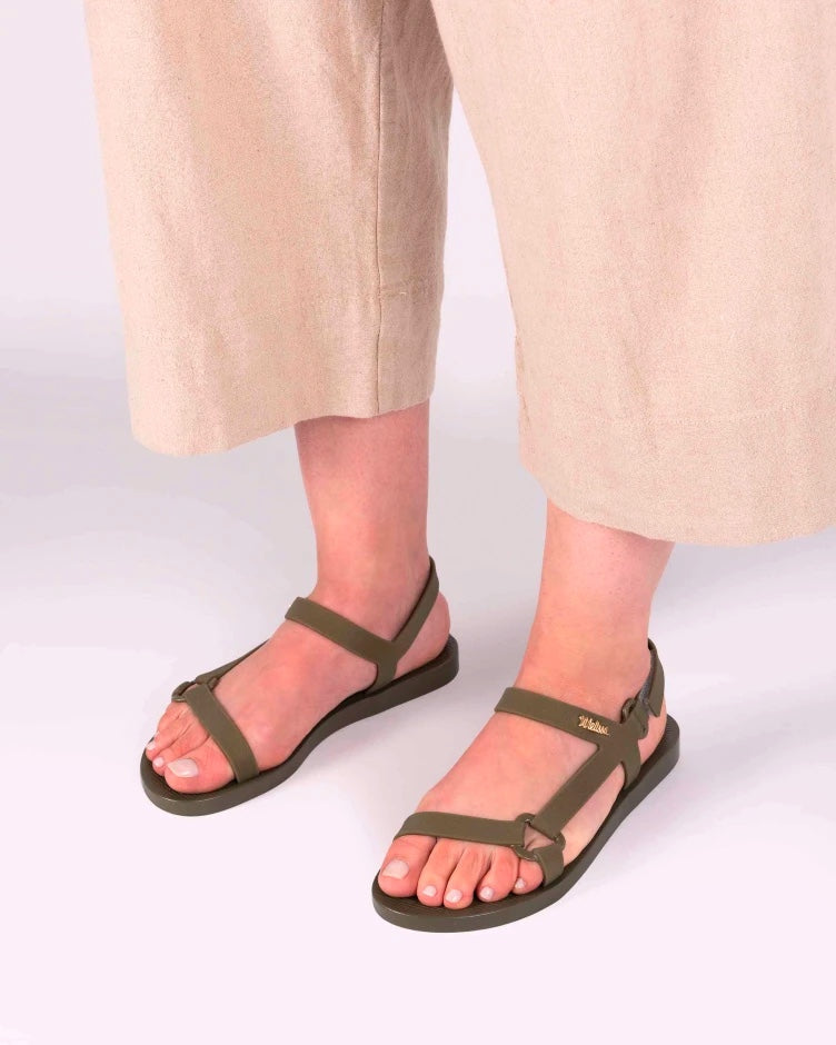 A model's legs wearing beige pants and a pair of green metallic Melissa Sun Downtown sandals with a front and ankle strap joined together by a vertical strap and a triangle buckle.