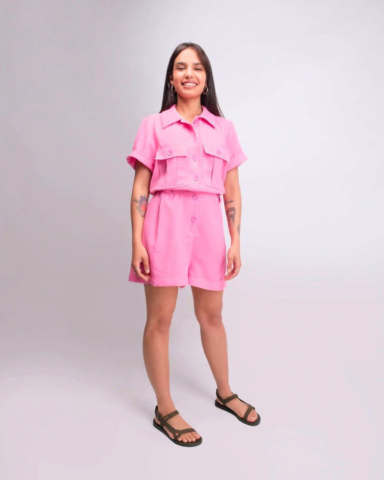 A model wearing a pink jumpsuit and a pair of green metallic Melissa Sun Downtown sandals with a front and ankle strap joined together by a vertical strap and a triangle buckle.