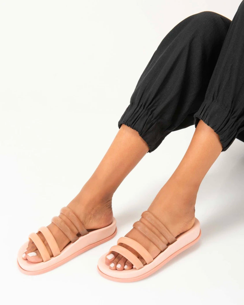 A model's legs in black pants and a pair of pink/clear pink Melissa Soft Wave Slides with 4 straps: two pink and two clear pink.